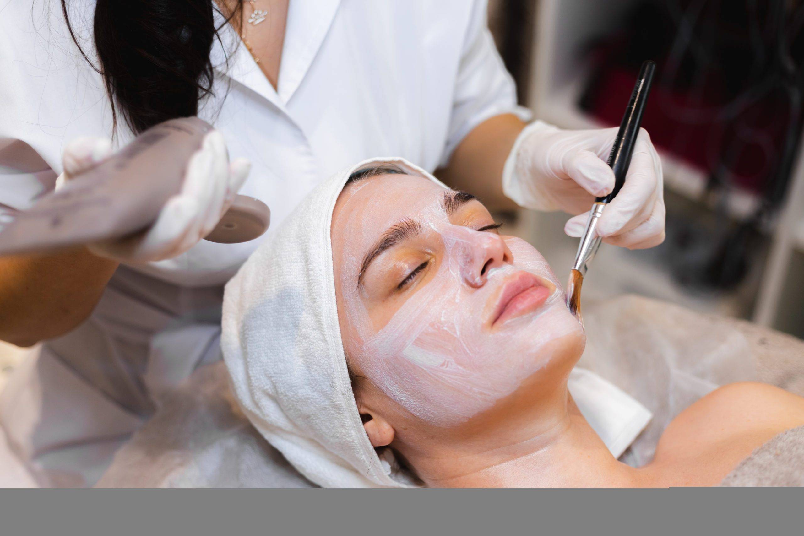 beautician-with-brush-applies-white-moisturizing-mask-face-young-girl-client-spa-beauty-salon-scaled